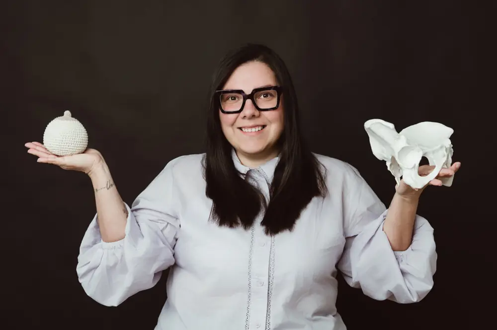 Katherine (Ka) Monge, doula y asesora de lactancia, sonriendo con un pecho tejido y una pelvis en las manos